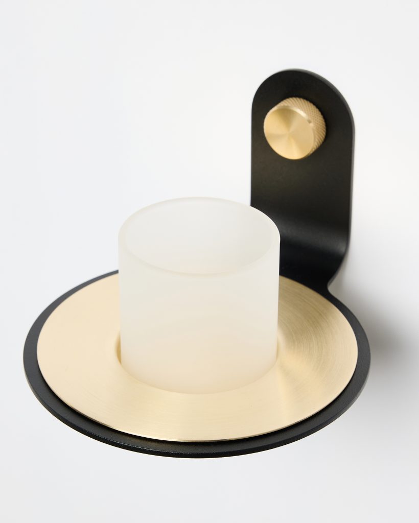 Primer plano de un producto WoodUpp: aplique portavelas de pared con cilindro de vidrio esmerilado, disco metálico dorado cepillado y soporte negro mate. Detalle del tornillo o pomo knurled dorado que sujeta la pieza, diseño minimalista moderno y acabado elegante.