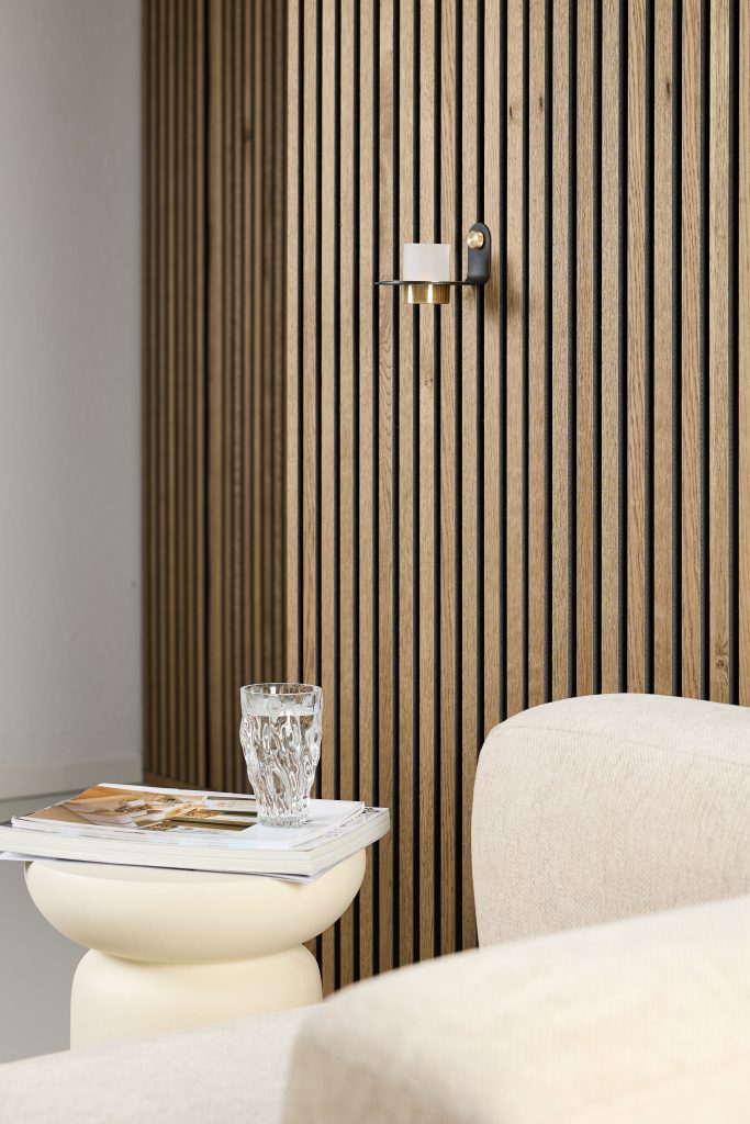 Interior minimalista con paneles verticales de listones de madera WoodUpp en tono roble; un pequeño aplique de pared negro con detalle dorado sostiene una vela, junto a un sofá beige, una mesa auxiliar crema con revistas y un vaso de agua sobre ellas.