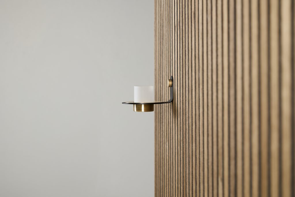 Panel de listones de madera WoodUpp en tono roble claro, instalado verticalmente, junto a un portavelas de pared WoodUpp: soporte metálico negro con detalle en latón, copa opalina para vela. Composición minimalista centrada en texturas y contrastes entre madera, metal y vidrio.