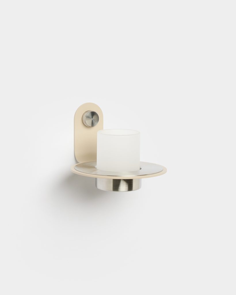 Imagen de WoodUpp que muestra un aplique de pared minimalista: placa trasera beige con botón metálico, plato circular y soporte de acero cepillado, y una tulipa cilíndrica de vidrio esmerilado blanca colocada sobre el portavelas. Diseño moderno, líneas limpias y acabados en metal y esmalte.