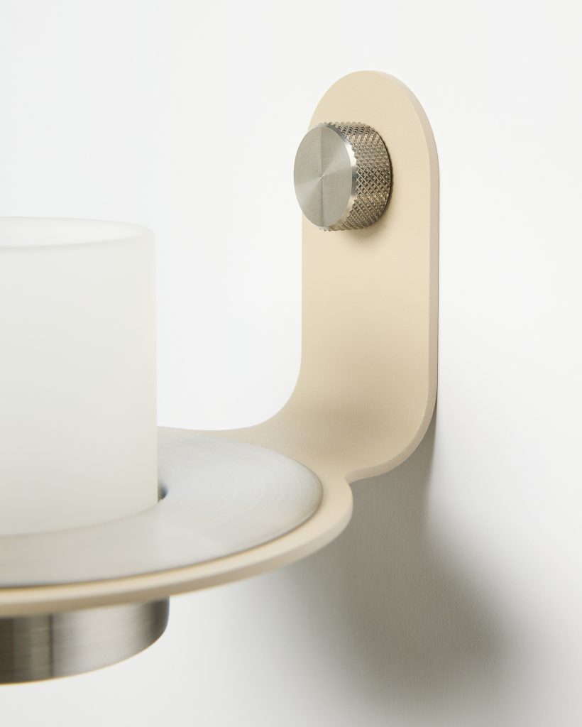 Primer plano de un aplique mural WoodUpp: soporte beige curvado con un tornillo metálico moleteado, plato circular con anillo cepillado y portavela de vidrio esmerilado en tonalidad blanca. Diseño minimalista y detalles metálicos visibles en una composición limpia contra pared blanca.