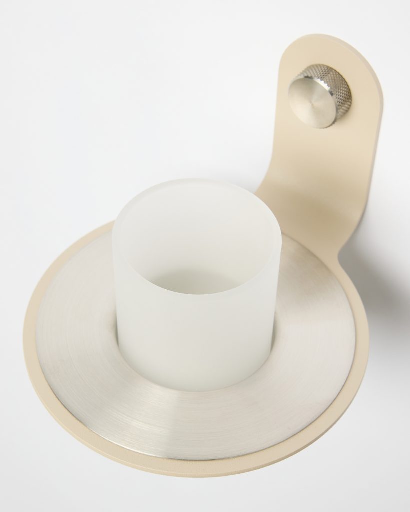 Imagen del accesorio WoodUpp: soporte mural beige con tornillo de ajuste metálico acanalado, anillo circular de acero cepillado y vaso cilíndrico de vidrio esmerilado blanco insertado, diseñado como portavasos/portacepillos sobrio y minimalista. Acabado mate, perfil curvo y tamaño compacto, ideal para baño moderno.