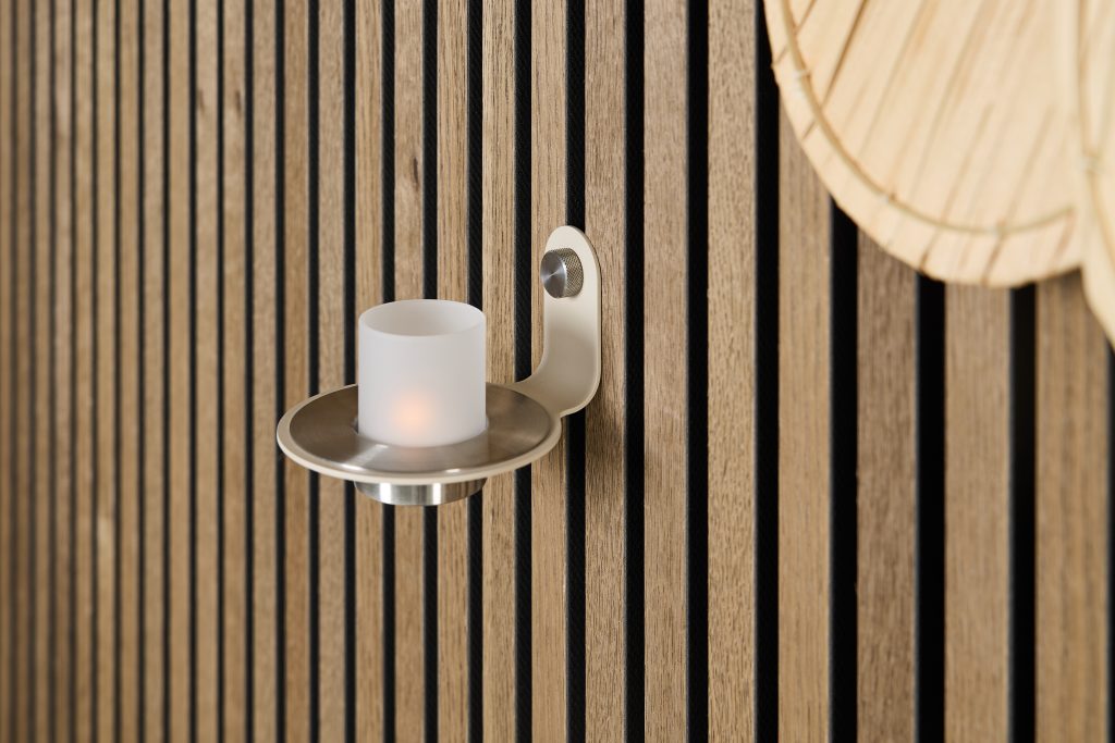 Paneles acanalados de madera WoodUpp en tono roble con ranuras negras verticales; soporte metálico WoodUpp montado en la lama sostiene una vela LED en vaso de vidrio esmerilado y una base circular de acero cepillado. Detalle lateral de adorno de fibra natural parcialmente visible.