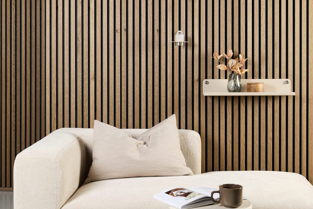 Interior minimalista con paneles de lamas de madera WoodUpp en vertical (vetas claras y ranuras negras), estantería flotante WoodUpp beige con tornillos visibles sosteniendo un jarrón de vidrio con hojas secas y una caja de madera; lámpara de pared, sofá y cojín crema en primer plano.